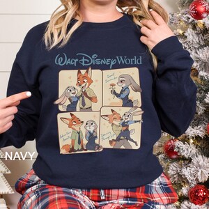 May include: Navy blue sweatshirt featuring a four-panel design with characters from the Disney movie Zootopia. The top panel has the text "Walt Disney World". The panels show Nick Wilde and Judy Hopps in various scenes. The word "NAVY" is printed on the bottom left.