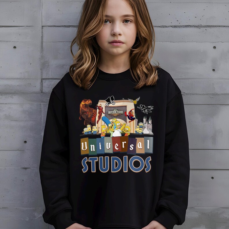 May include: Black sweatshirt featuring a colorful graphic of Universal Studios characters, including dinosaurs, Minions, and The Simpsons. The text "Universal Studios" is displayed in a multi-colored block design. The sweatshirt is worn by a person.