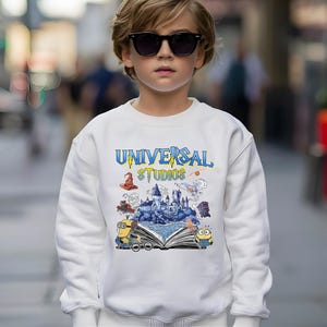 Op de afbeelding: Een witte sweatshirt met de tekst "UNIVERSAL STUDIOS" in blauw en geel. De afbeelding bevat een kasteel, Minions en andere themaparkelementen. De sweatshirt wordt gedragen door een persoon met een zonnebril.