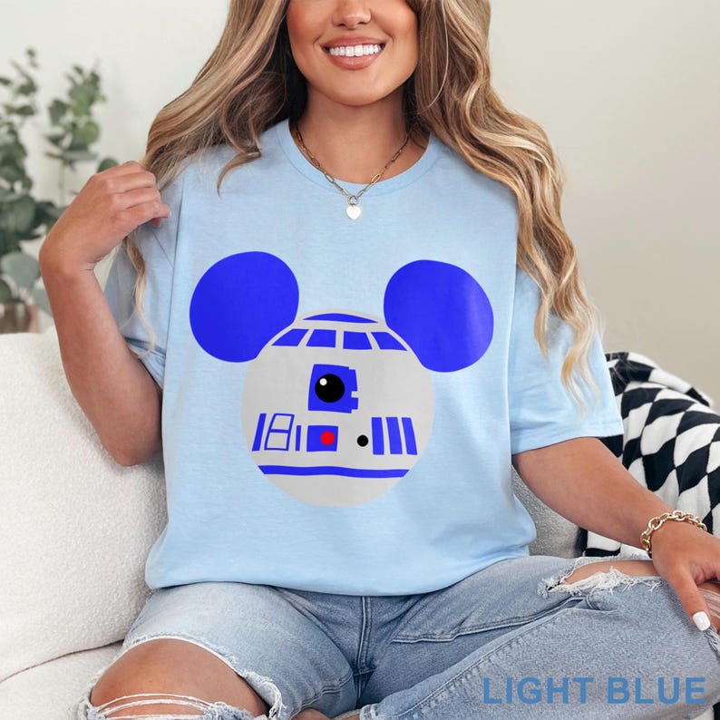 May include: Light blue t-shirt featuring a graphic of a droid with Mickey Mouse ears. The droid design is white and blue, with a red and black detail. The text "LIGHT BLUE" is at the bottom of the image.