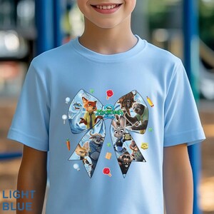 Op de afbeelding: Lichtblauw T-shirt met een kleurrijke afbeelding van personages uit de animatiefilm "Zootopia 2" in de vorm van een strik. De tekst "Zootopia 2" is zichtbaar. Het shirt heeft ook de tekst "LIGHT BLUE" onderaan.