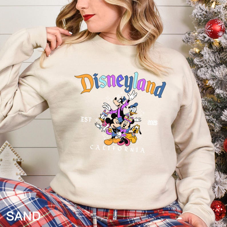 Puede incluir: Una sudadera de color arena con la palabra "Disneyland" en letras coloridas y un gr&aacute;fico de personajes de Disney. El gr&aacute;fico incluye Mickey Mouse, Minnie Mouse, Donald Duck, Goofy y Pluto. Tambi&eacute;n es visible el texto "EST 2023 CALIFORNIA".