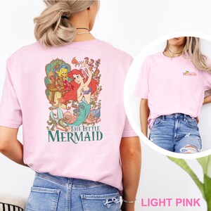 May include: A light pink t-shirt featuring a large graphic on the back of Ariel and Flounder from The Little Mermaid surrounded by floral elements and the text The Little Mermaid. The front chest area displays a small The Little Mermaid logo.