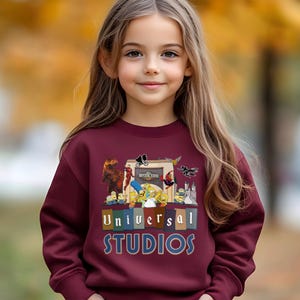 May include: A maroon sweatshirt featuring the Universal Studios logo and various cartoon characters. The design includes images of dinosaurs, Minions, The Simpsons, and Spider-Man. The sweatshirt is worn by a young girl with long brown hair.