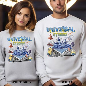 Op de afbeelding: Sportgrijze en witte sweatshirts met het "Universal Studios" logo in blauw en geel. Het ontwerp bevat een kasteel, Minions en andere graphics. De sweatshirts worden getoond op een vrouw en een man.