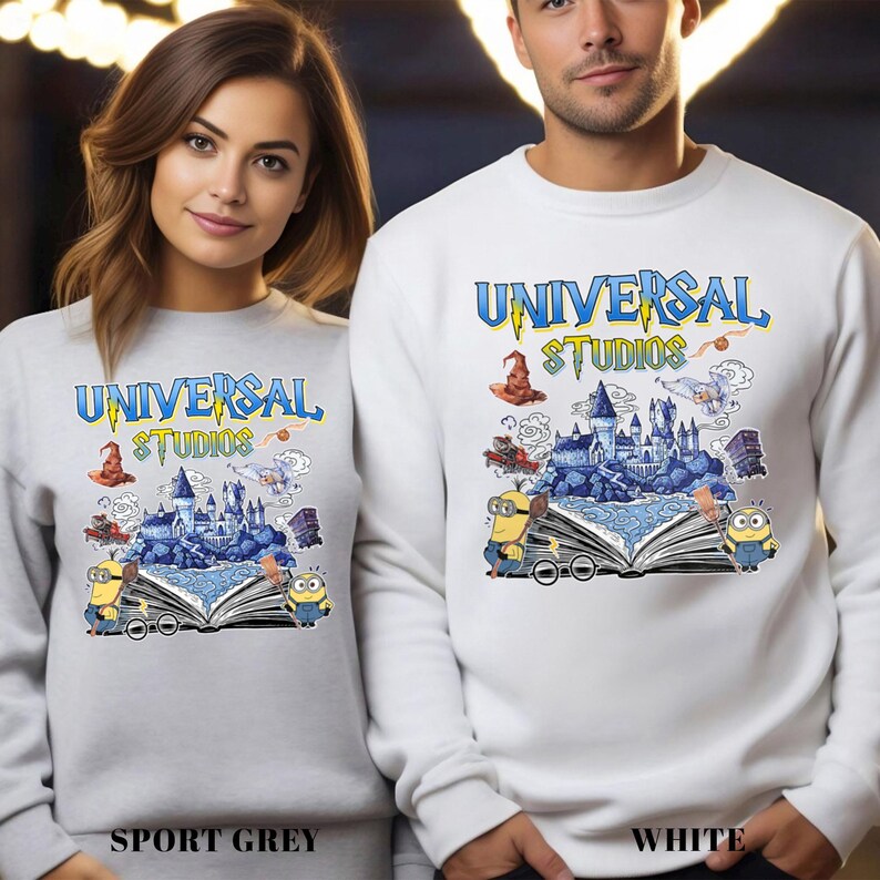 Op de afbeelding: Sportgrijze en witte sweatshirts met het "Universal Studios" logo in blauw en geel. Het ontwerp bevat een kasteel, Minions en andere graphics. De sweatshirts worden getoond op een vrouw en een man.