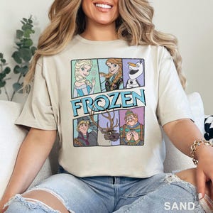May include: Sand-colored t-shirt featuring a graphic of characters from the movie "Frozen." The design includes Elsa, Anna, Olaf, Kristoff, Sven, and a character with a striped shirt. The word "FROZEN" is in a blue and white font.