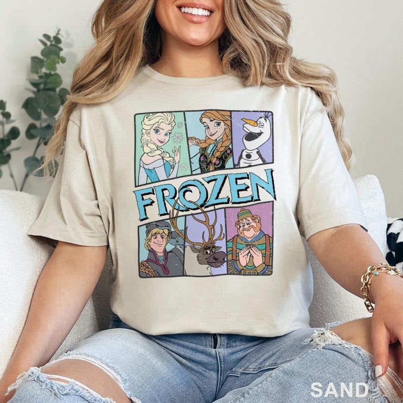 May include: Sand-colored t-shirt featuring a graphic of characters from the movie "Frozen." The design includes Elsa, Anna, Olaf, Kristoff, Sven, and a character with a striped shirt. The word "FROZEN" is in a blue and white font.
