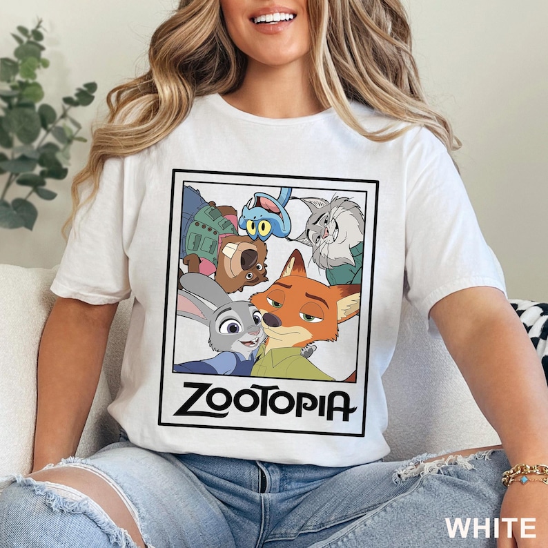 May include: White t-shirt featuring a graphic from the animated movie Zootopia. The graphic includes characters like Judy Hopps and Nick Wilde. The word "Zootopia" is printed in a bold, black font. The shirt is made of a soft material.