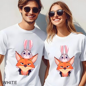 May include: White t-shirts featuring a cartoon fox and bunny design. The fox is orange and the bunny is gray with pink ears. The word "WHITE" is printed at the bottom of the shirt. The shirts are worn by two people.