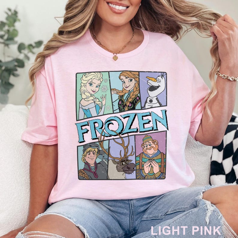 May include: Light pink t-shirt featuring a graphic of characters from the movie "Frozen". The design includes Elsa, Anna, Olaf, Kristoff, Sven, and a character in a blue hat. The word "FROZEN" is printed in large, blue letters.