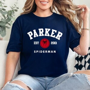 May include: Navy blue t-shirt featuring the name "PARKER" in white, arched above a red circle with a spider symbol and the text "EST 2001". Below, the word "SPIDERMAN" is printed in white. The shirt is a casual wear item.
