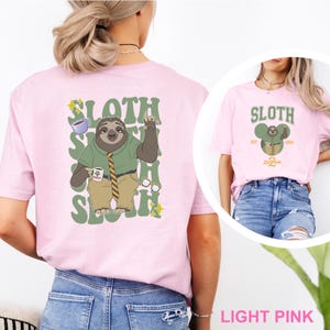 May include: Light pink t-shirt featuring a sloth character design. The back of the shirt has a large graphic of a sloth in a green shirt, tie, and khaki pants, holding a coffee mug. The word "SLOTH" is printed in green. The front of the shirt has a smaller sloth design.