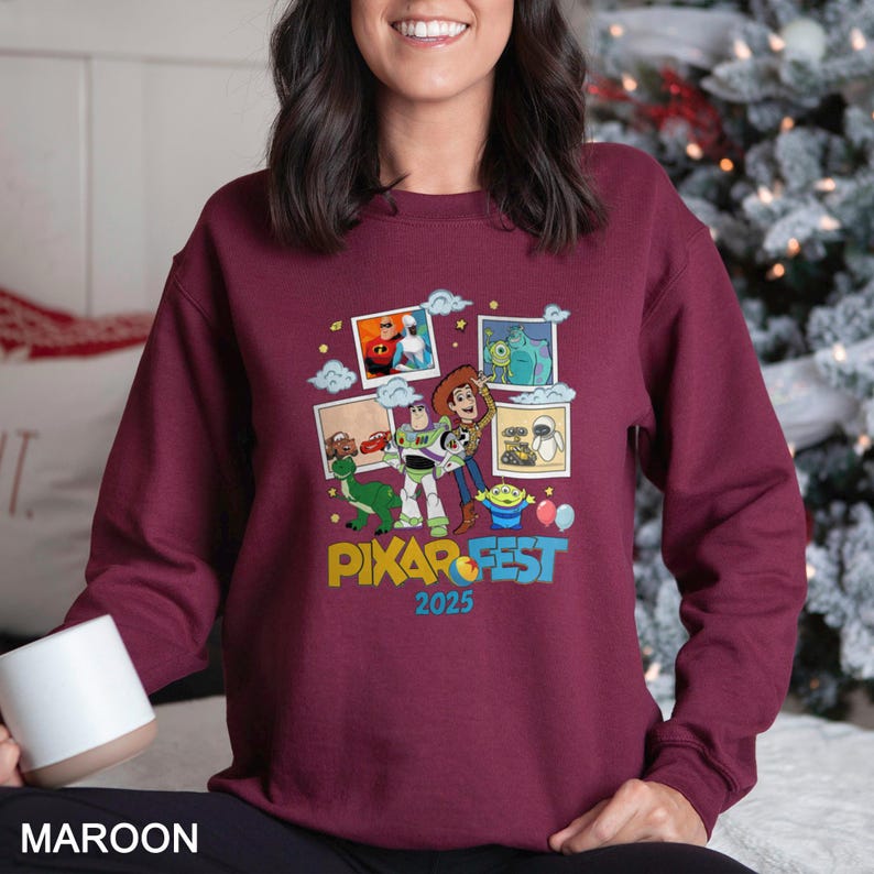 Może przedstawiać: Bordowa bluza z kolorową grafiką postaci z filmu Pixar i napisem "PIXAR FEST 2025". Projekt zawiera obrazy postaci takich jak Buzz Lightyear i Woody. Bluza jest w jednolitym bordowym kolorze.