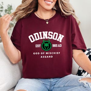 May include: A maroon t-shirt with the word "ODINSON" in white block letters. Below the word is a green and black graphic and the text "EST 965 AD". The shirt also has the text "GOD OF MISCHIEF ASGARD".