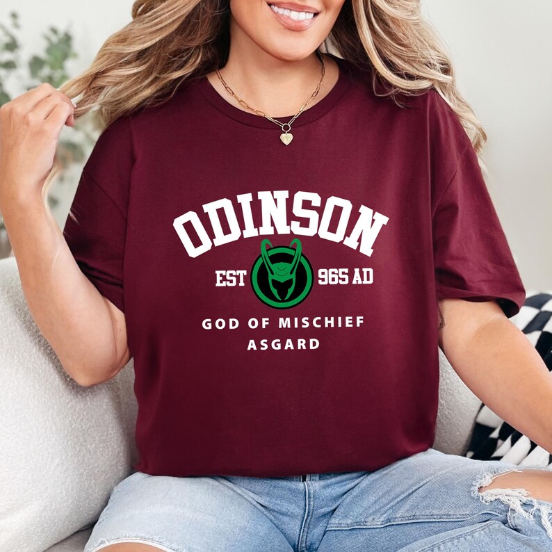 May include: A maroon t-shirt with the word "ODINSON" in white block letters. Below the word is a green and black graphic and the text "EST 965 AD". The shirt also has the text "GOD OF MISCHIEF ASGARD".