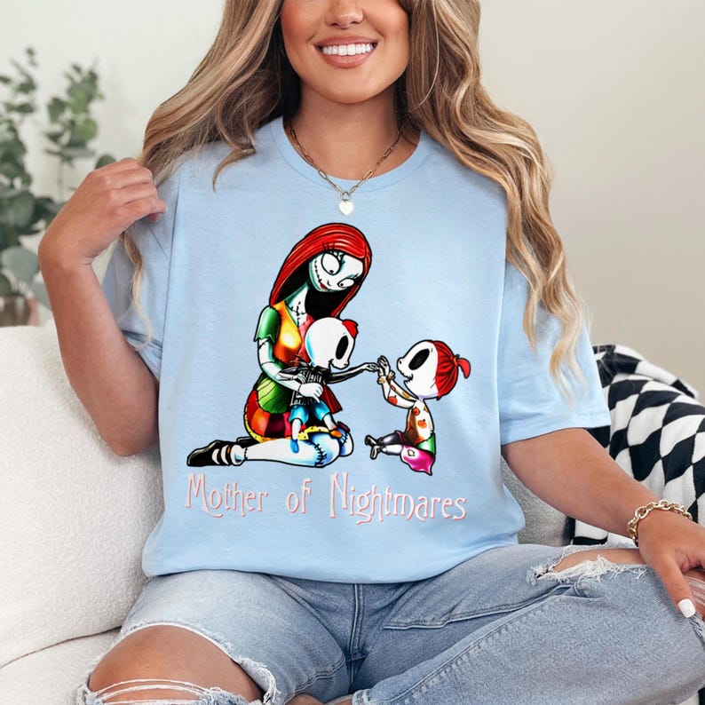 Puede incluir: Camiseta azul claro con un gr&aacute;fico colorido de un personaje con cabello rojo y un vestido de retazos, con dos ni&ntilde;os esqueleto. El texto "Mother of Nightmares" est&aacute; impreso debajo. La camiseta es de material suave.