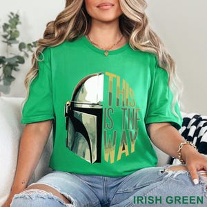 Pu&ograve; includere: Una t-shirt a maniche corte di colore verde irlandese brillante con una grafica di un casco mandaloriano argentato sul lato sinistro e il testo THIS IS THE WAY in un carattere a blocchi impilato sul lato destro, in toni oliva e verde giallo.
