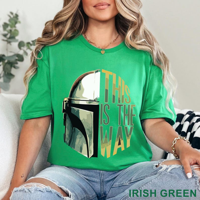 Pu&ograve; includere: Una t-shirt a maniche corte di colore verde irlandese brillante con una grafica di un casco mandaloriano argentato sul lato sinistro e il testo THIS IS THE WAY in un carattere a blocchi impilato sul lato destro, in toni oliva e verde giallo.