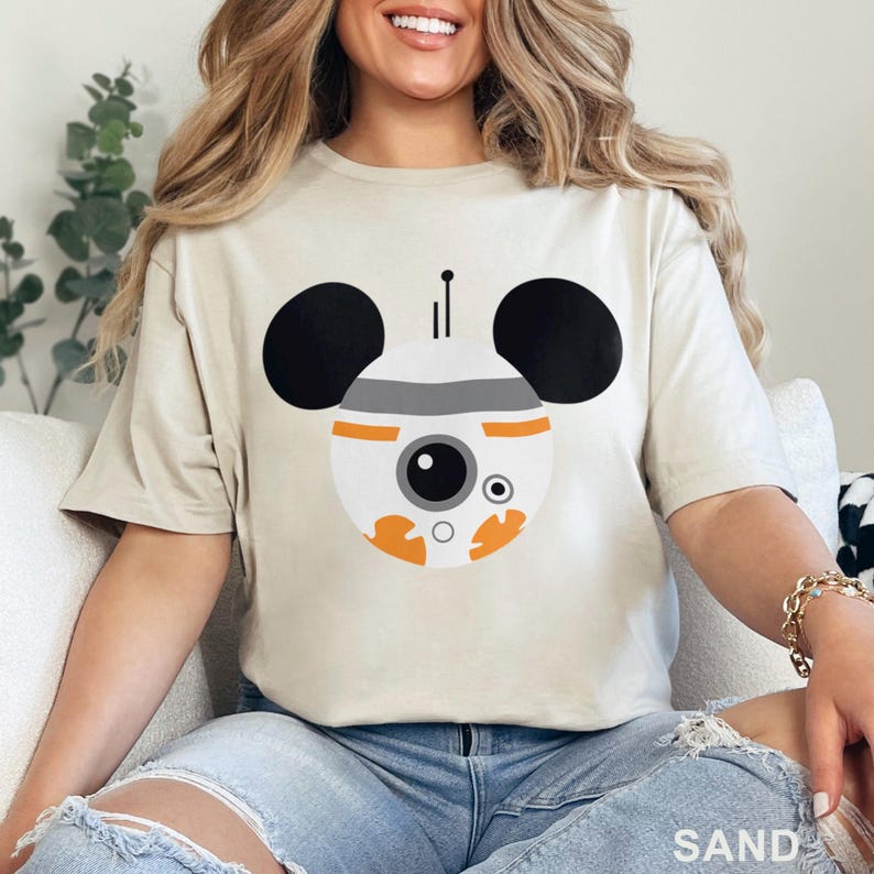 May include: Sand-colored t-shirt featuring a BB-8 inspired design with Mickey Mouse ears. The graphic includes a white and orange droid face with black ears, a gray stripe, and a black antenna. The word "SAND" is printed at the bottom.