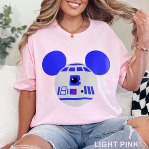 May include: Light pink t-shirt featuring a graphic of R2-D2 as Mickey Mouse. The design includes blue ears, a gray head, and blue and red details. The text "LIGHT PINK" is at the bottom of the image.