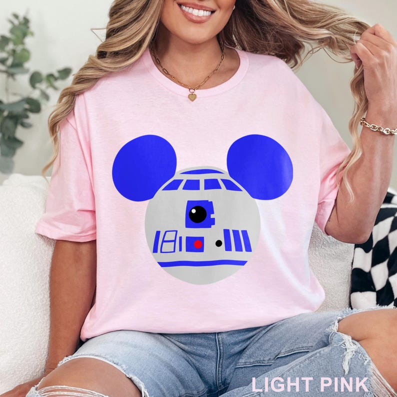 May include: Light pink t-shirt featuring a graphic of R2-D2 as Mickey Mouse. The design includes blue ears, a gray head, and blue and red details. The text "LIGHT PINK" is at the bottom of the image.