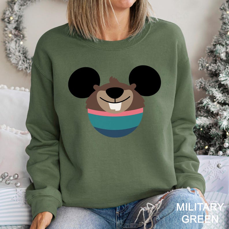 May include: Military green sweatshirt featuring a cartoon character design. The design includes a brown face with black ears, a pink and teal striped circle, and a single white tooth. The text "MILITARY GREEN" is printed on the bottom right.
