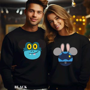 May include: Black sweatshirts with cartoon character designs. One features a blue monster face with yellow eyes, the other a police officer design. The word "BLACK" is printed at the bottom. The sweatshirts are worn by a man and a woman.
