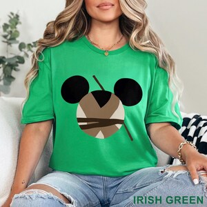May include: A green t-shirt with a graphic design featuring a stylized face with black ears, a brown and beige center, and two brown lines. The text "IRISH GREEN" is at the bottom of the image.