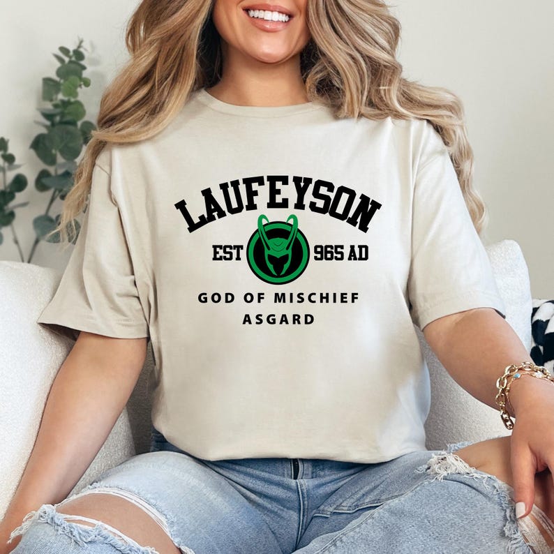 May include: Beige t-shirt with the word "LAUFEYSON" in black, above a green and black emblem. Below the emblem are the words "EST 965 AD", "GOD OF MISCHIEF", and "ASGARD" in black. The shirt is worn by a person.