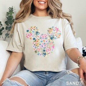 May include: Sand-colored t-shirt featuring a floral Mickey Mouse design. The design is made of various colorful flowers, including pink, blue, and yellow. The word "SAND" is printed at the bottom of the image.