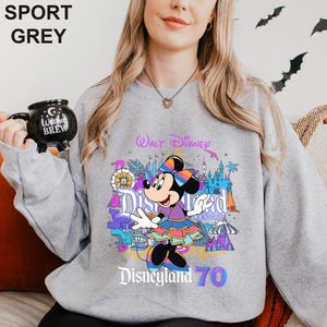 May include: Sport grey sweatshirt featuring a colorful graphic of Minnie Mouse in front of Disneyland Resort. The design includes the text "Walt Disney Disneyland 70". The sweatshirt is a light grey color.