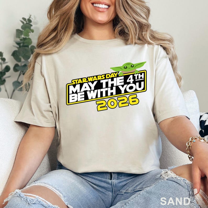 May include: Sand-colored t-shirt featuring the text "STAR WARS DAY MAY THE 4TH BE WITH YOU 2026" in yellow and white, with a green Baby Yoda graphic. The shirt is a casual style.