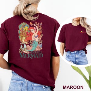 May include: A maroon t-shirt featuring a large graphic on the back of Ariel and Flounder from The Little Mermaid surrounded by floral elements and the text The Little Mermaid. The front chest area displays a small logo with the same movie title.