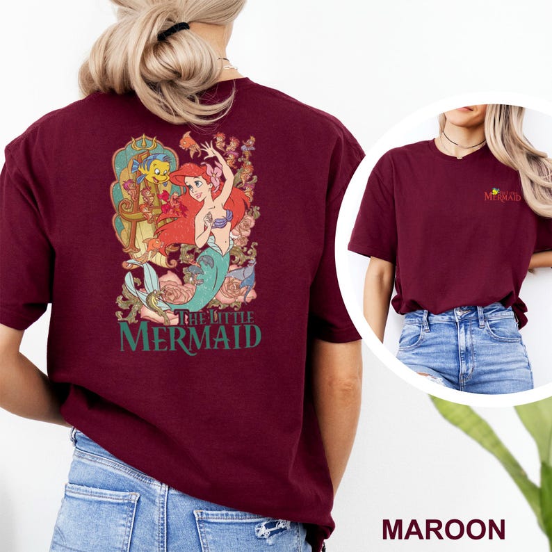 May include: A maroon t-shirt featuring a large graphic on the back of Ariel and Flounder from The Little Mermaid surrounded by floral elements and the text The Little Mermaid. The front chest area displays a small logo with the same movie title.