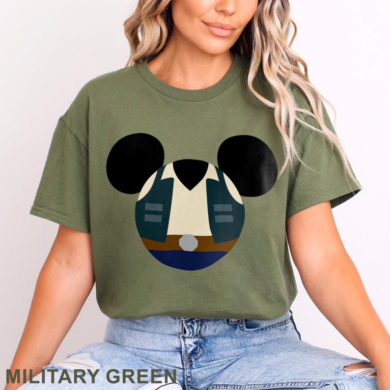 May include: Military green t-shirt featuring a cartoon design of a character's outfit within a Mickey Mouse head shape. The design includes a white shirt, a dark blue vest, and a brown belt. The text "MILITARY GREEN" is at the bottom.