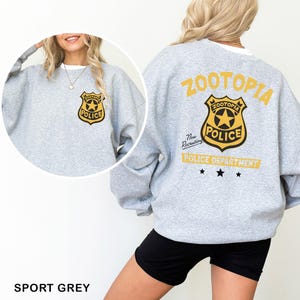May include: Sport grey sweatshirt featuring a Zootopia Police Department badge on the front and back. The back design includes the text "ZOOTOPIA POLICE DEPARTMENT" in yellow. The sweatshirt has long sleeves and a crew neck.