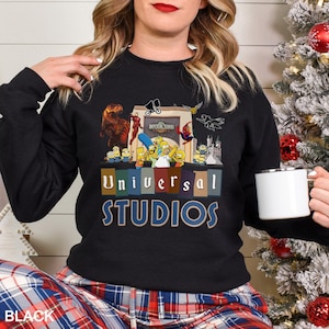 May include: Black crewneck sweatshirt featuring a colorful graphic of Universal Studios characters, including the Simpsons, Minions, and Spider-Man. The words "Universal Studios" are printed in blue and gold. The word "BLACK" is printed at the bottom.