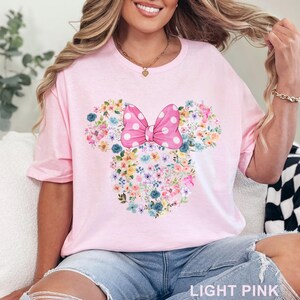 May include: Light pink t-shirt featuring a floral Minnie Mouse design. The design includes a pink polka dot bow and floral elements in various colors. The text "LIGHT PINK" is at the bottom of the shirt.