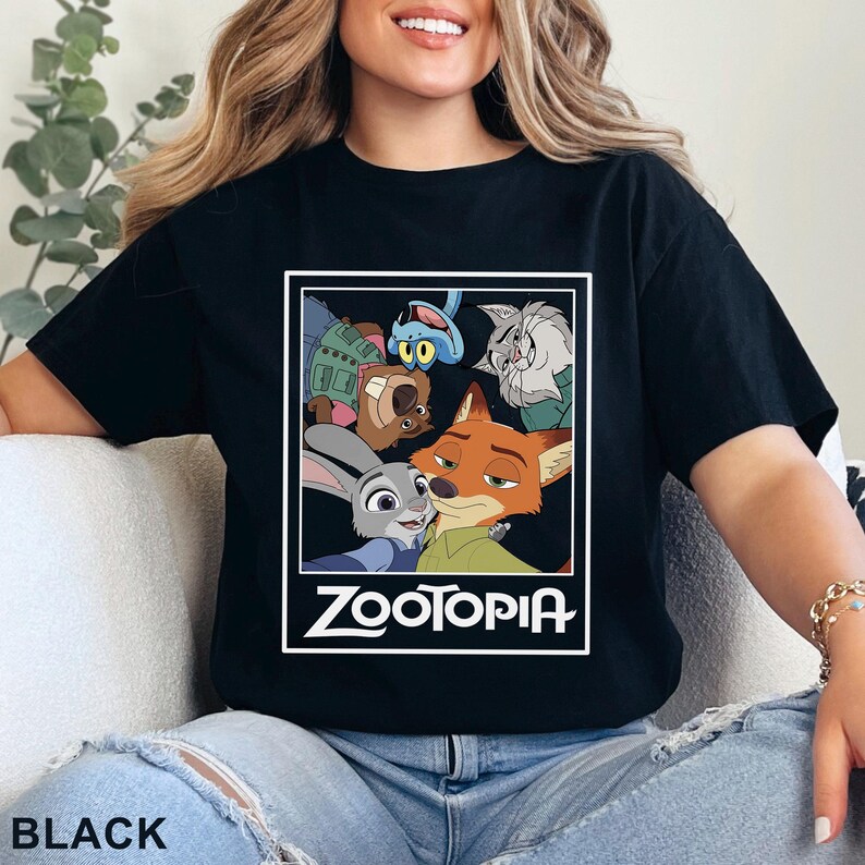May include: Black t-shirt with a rectangular graphic of characters from the animated film "Zootopia." The graphic includes Judy Hopps, Nick Wilde, and other characters. The word "ZOOTOPIA" is printed in white below the characters. The word "BLACK" is printed at the bottom.