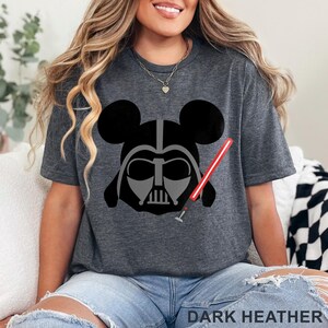 May include: Dark heather gray t-shirt featuring a graphic of Darth Vader's helmet combined with Mickey Mouse ears and a red lightsaber. The text "DARK HEATHER" is printed at the bottom of the shirt.