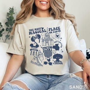May include: Sand-colored t-shirt with a navy blue graphic design featuring the text "The Most Magical Place on Earth" and various Disney-themed icons, including a castle, Mickey Mouse ears, and a smiling face.