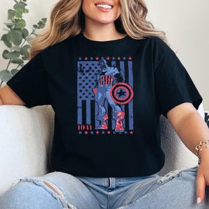 May include: Black t-shirt featuring a patriotic graphic of a superhero with a shield, set against a backdrop of the American flag. The design includes the year "1941" and stars. The shirt is a classic crew neck.