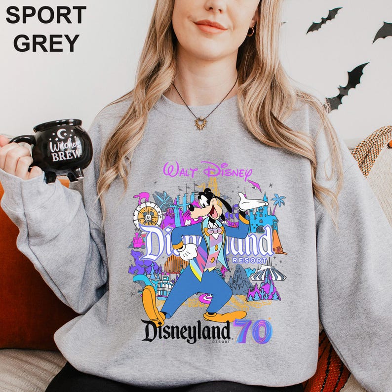 May include: Sport grey sweatshirt featuring a colorful graphic of Goofy from Disneyland Resort. The design includes the text "Walt Disney", "Disneyland Resort", and "Disneyland 70". The sweatshirt is a light grey color.