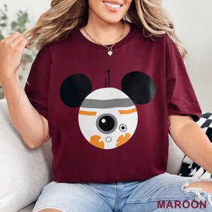 May include: Maroon t-shirt featuring a cartoon design of a droid with Mickey Mouse ears. The droid is white, gray, and orange with a large black eye. The word "MAROON" is printed at the bottom of the shirt.