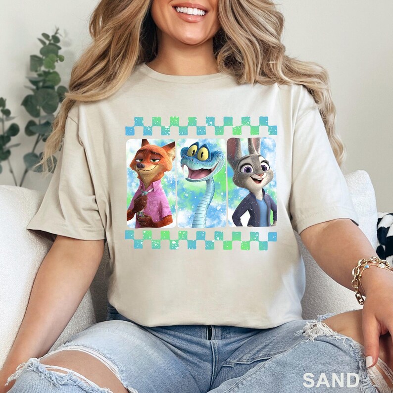 May include: Sand-colored t-shirt featuring three cartoon characters: a fox in a pink shirt, a blue snake, and a rabbit in a blue jacket. The characters are set against a blue and green background within a checkered border.