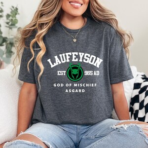 May include: Dark gray t-shirt with the text "LAUFEYSON" above a green and black graphic and the words "EST 965 AD". Below the graphic are the words "GOD OF MISCHIEF ASGARD". The shirt has a crew neck.