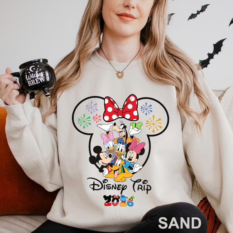 May include: Sand-colored sweatshirt featuring a large graphic of Disney characters, including Mickey and Minnie Mouse, Donald and Daisy Duck, and Goofy. The design includes a red bow with white polka dots and the text "Disney Trip 2023".