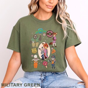 K&ouml;nnte beinhalten: Ein milit&auml;rgr&uuml;nes T-Shirt mit einem Zootopia-Grafikdruck. Das Design zeigt Charaktere wie Judy Hopps und Nick Wilde, eine Polizeimarke, einen Donut, eine Karotte und ein pfotenf&ouml;rmiges Eis am Stiel, mit dem Zootopia-Logo oben in der Mitte.