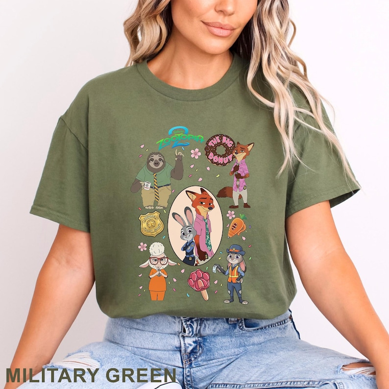 K&ouml;nnte beinhalten: Ein milit&auml;rgr&uuml;nes T-Shirt mit einem Zootopia-Grafikdruck. Das Design zeigt Charaktere wie Judy Hopps und Nick Wilde, eine Polizeimarke, einen Donut, eine Karotte und ein pfotenf&ouml;rmiges Eis am Stiel, mit dem Zootopia-Logo oben in der Mitte.